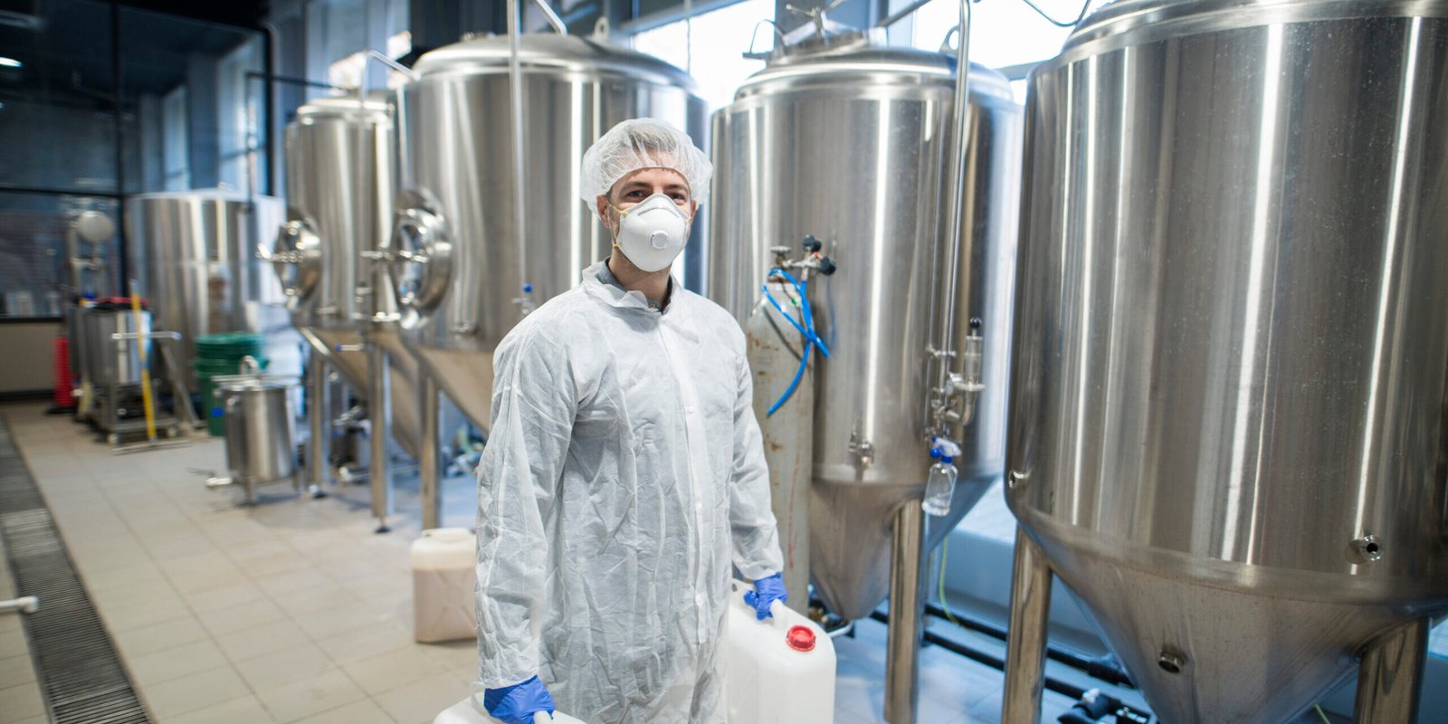 Industrial worker technologist in white protective suit with hairnet and mask holding plastic cans with chemicals in food factory production line. Industrial worker technologist in white protective suit with hairnet and mask holding plastic cans with chemicals in food factory production line.